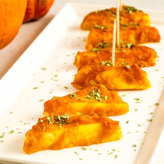 A serving plate with baked paneer in pumpkin sauce with sticks attached to each piece.