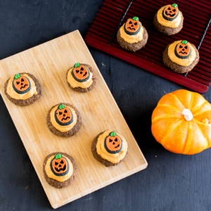 Top view of frosted Pumpkin Cheesecake Coffee Flax Cookies on a wooden board and a red mat on the side with a pumpkin as the prop