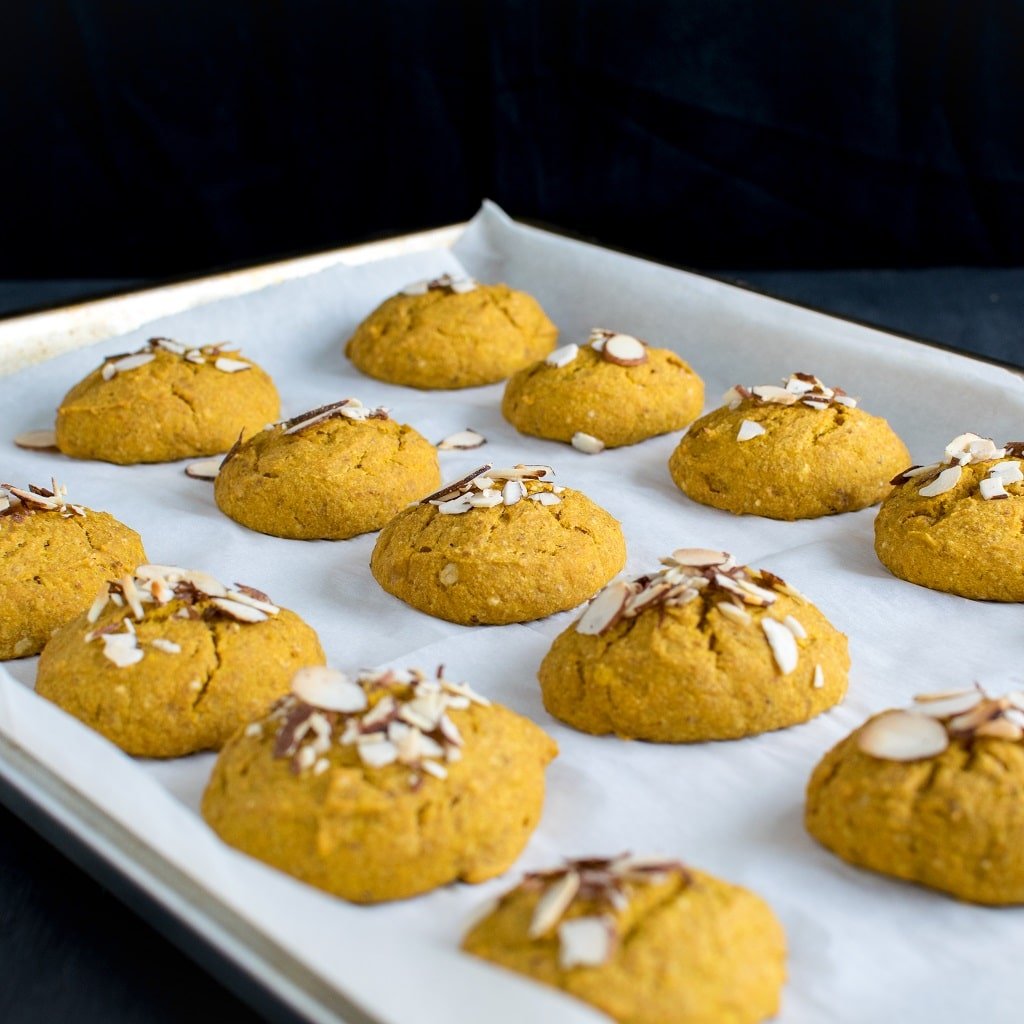 Almond Butter Pumpkin Oatmeal Cookies are shown on a cookie sheet fresh out of the oven. The cookie sheet is covered with a parchment paper and the cookies are beautifully spread over it .
