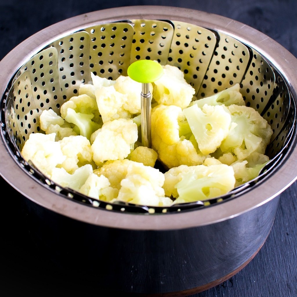 Steamed cauliflower florets are shown in a steamer basket placed inside a saucepan 