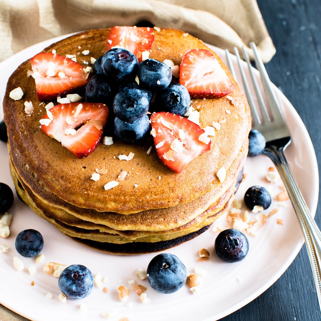 A pink plate stacked with chickpea flour peanut butter protein pancakes topped with fruits and nuts is shown in this image