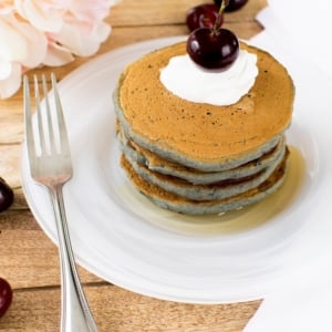 A stack of Cherry Oatmeal Vegan pancakes are shown in a wound white marble plate with whipped cream and a cherry on top