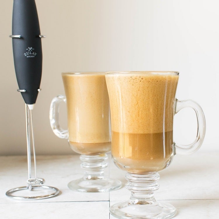 Two tall glasses filled with iced cappuccino and milk frother on the side
