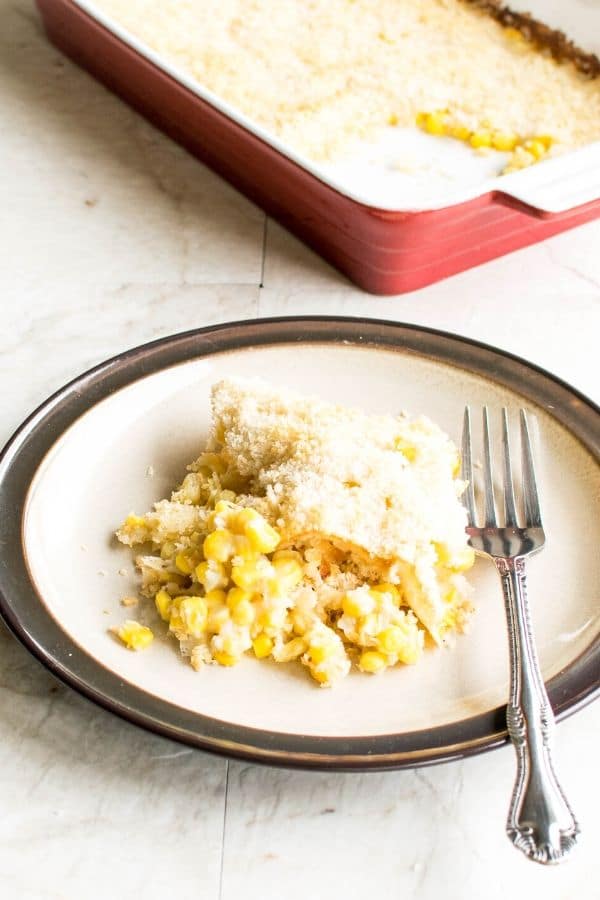 A portion of creamed corn casserole on a serving plate with the entire casserole dish at the background