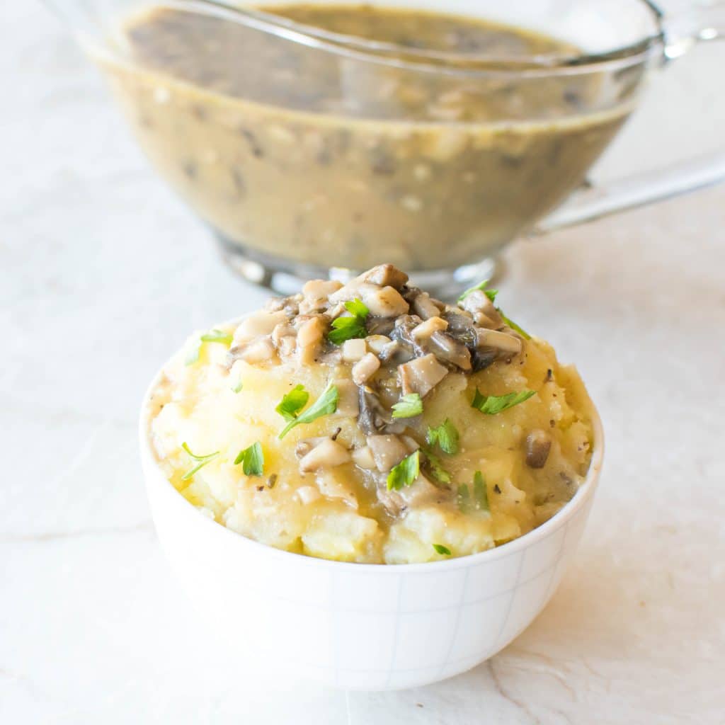 A bowl loaded with mashed potatoes and topped with vegan gravy. A jar filled with vegan gravy at the background.