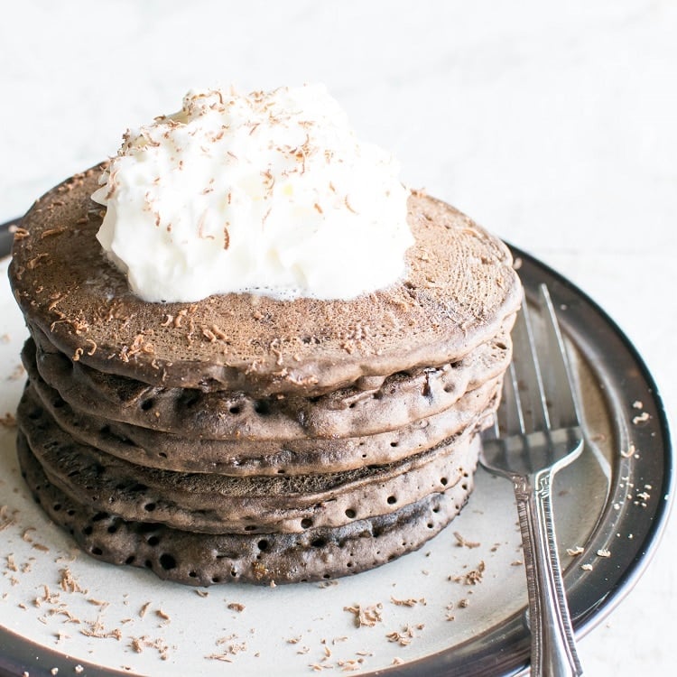 A close up front view of a stack of vegan hot chocolate buckwheat pancakes with toppings and a fork on the side
