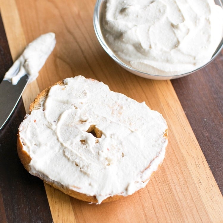 A close up view of a bagel with vegan cream cheese spread and a butter knife on the side on a wooden board