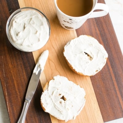 Bagels with Nut Free Vegan Cream Cheese spread on them along with a bowl full of the dip and a cup of coffee on a wooden board