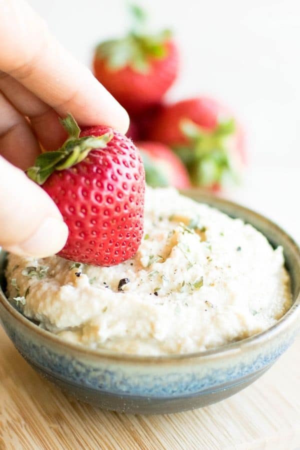 A hand dipping strawberry into vegan ricotta cheese