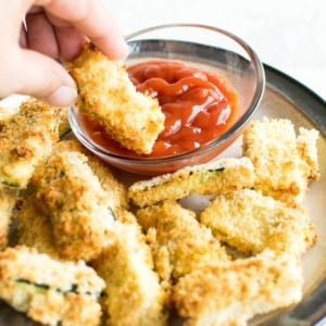A hand dipping air fryer zucchini fries into ketchup