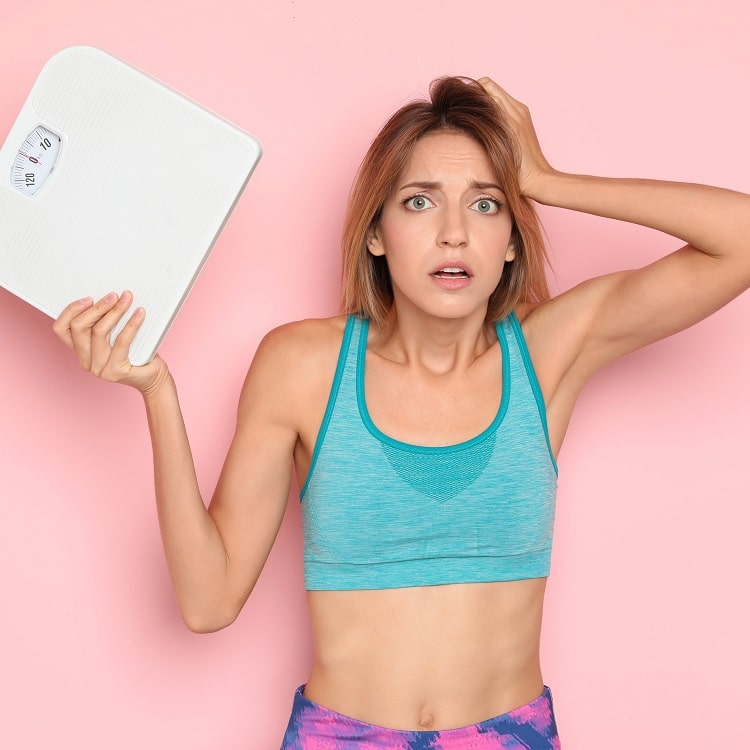 A woman holding a bathroom weighing scale and showing what happens when we restrict calories.