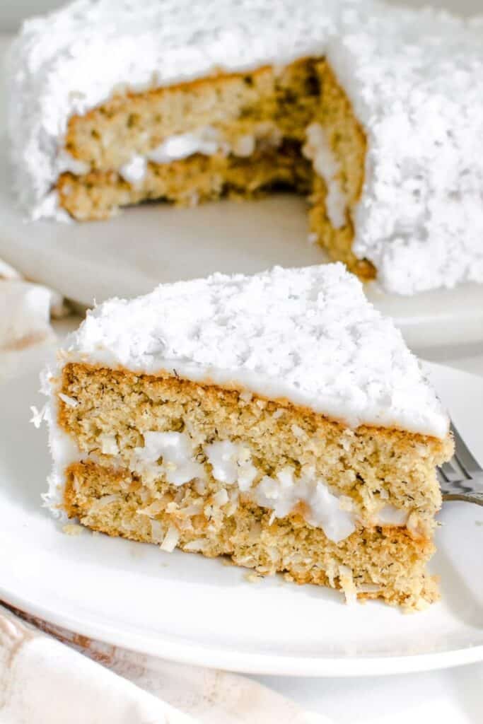 a slice of vegan coconut cake on a serving plate with the entire cake at the backdrop.