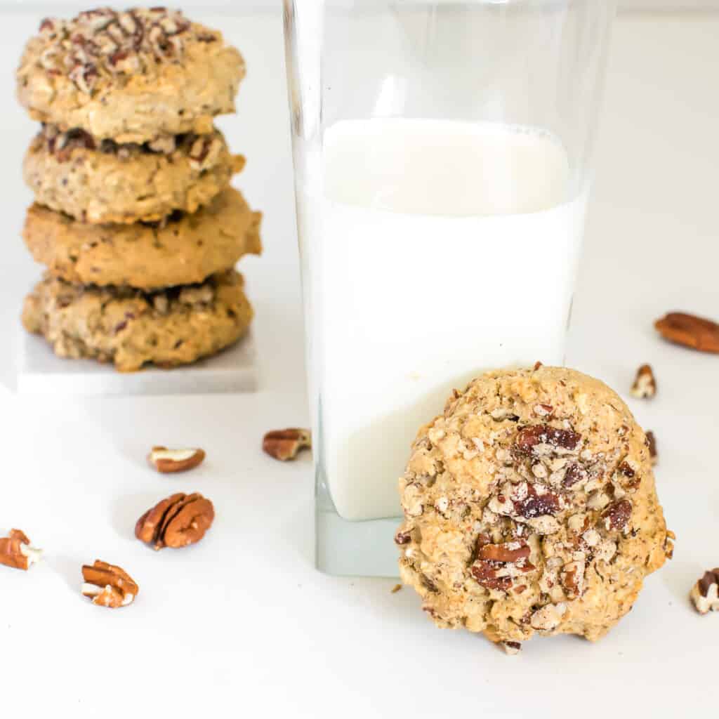 focus on a vegan pecan cookie with a glass of milk and some stacked at the background.